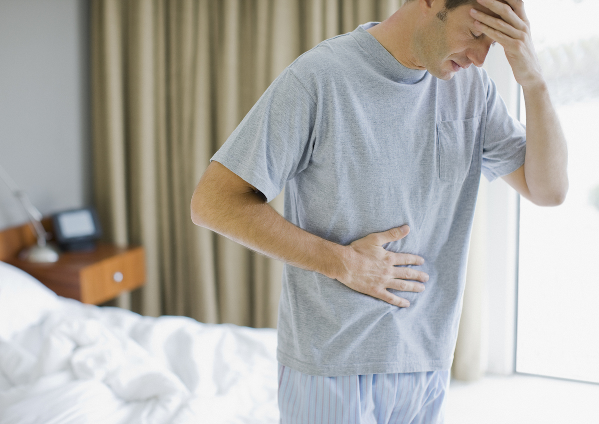 A man is standing beside a bed and holding his head and abdomen in his hands.