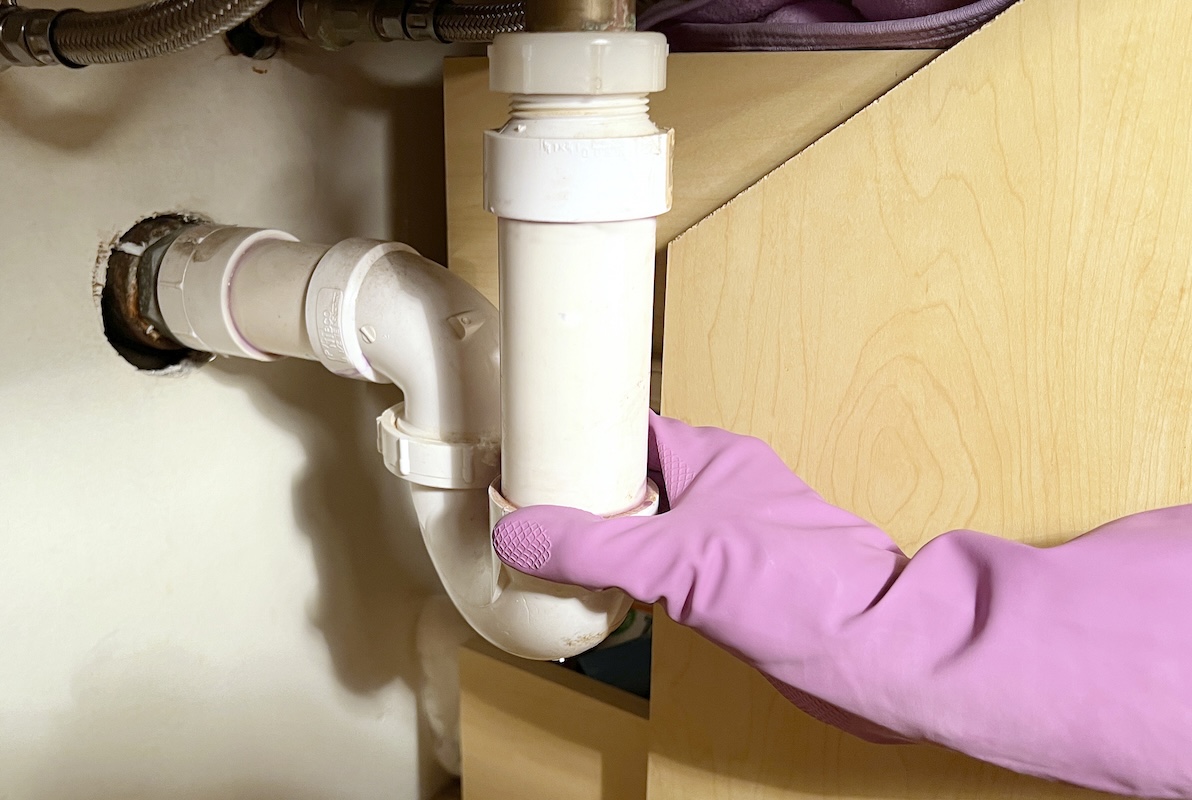 Person wearing rubber cleaning gloves inspects pipes under sink.