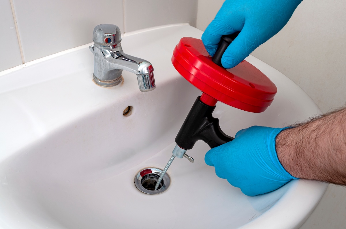 Person wearing blue gloves puts drain auger down a white bathroom sink.