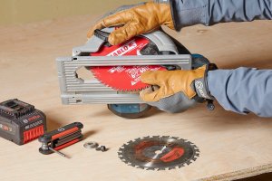 Gloved hands hold a circular saw, swapping a new blade into the saw—the alternate blade is on the table next to a tool battery and hex wrench.