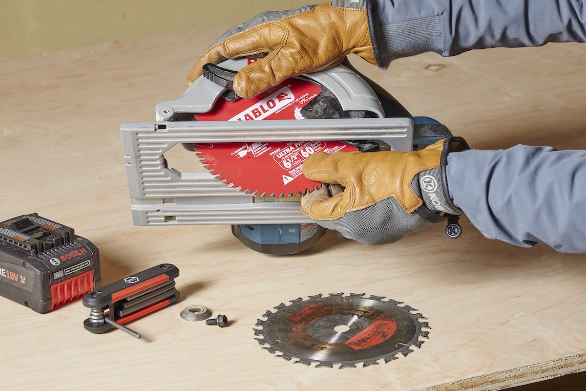 Gloved hands hold a circular saw, swapping a new blade into the saw—the alternate blade is on the table next to a tool battery and hex wrench.
