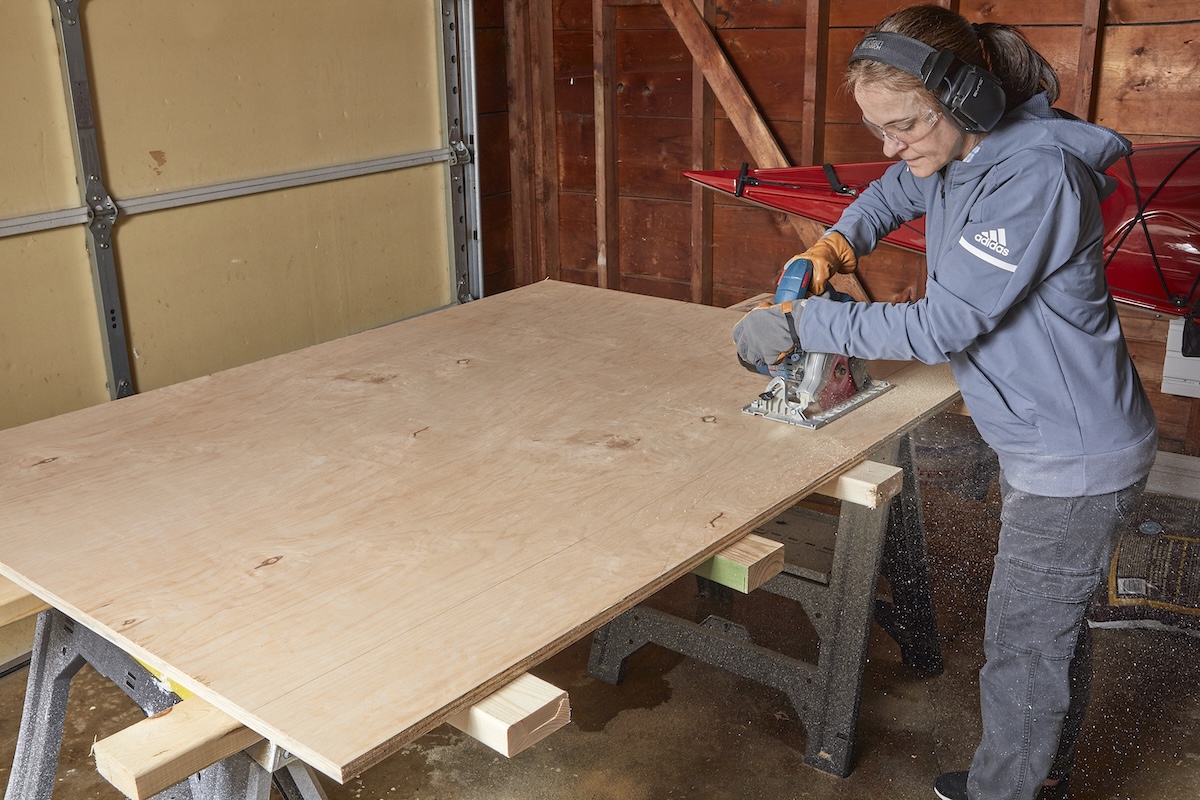 Woman wearing protective eyewear and ear coverings uses a circular saw to make a rip cut in plywood.