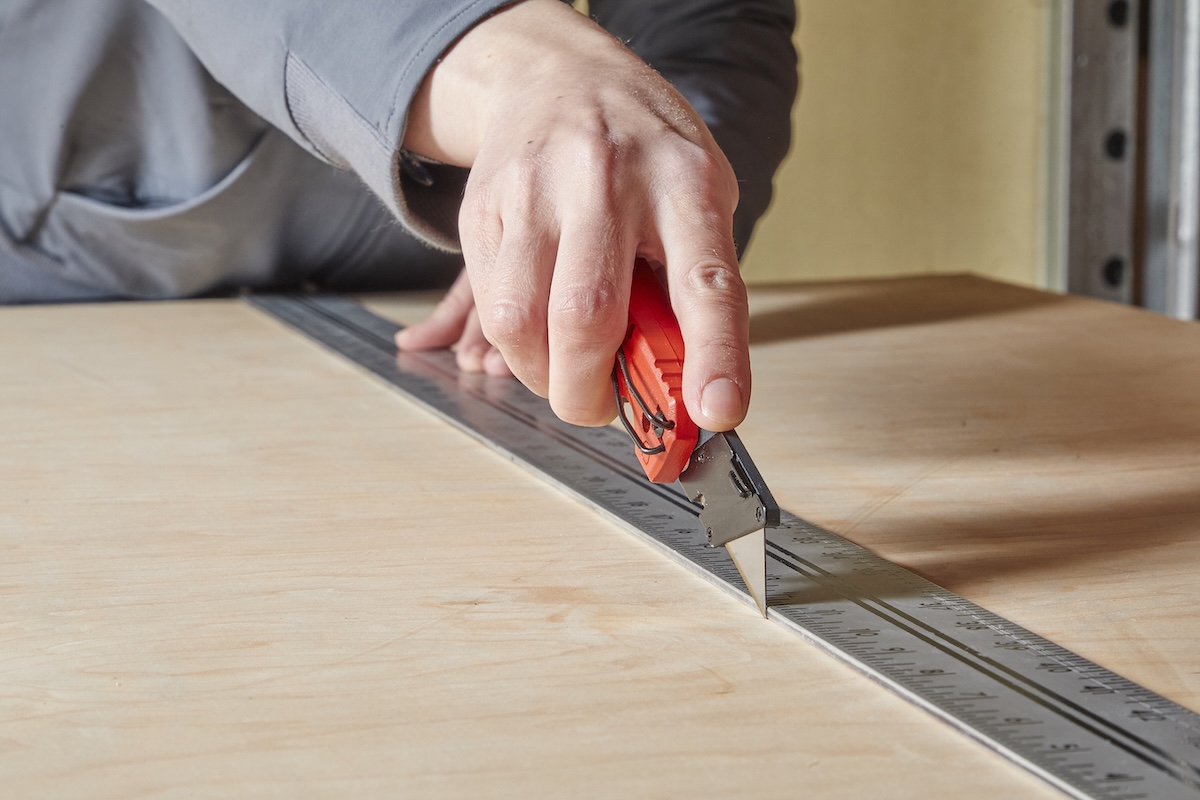 Woman uses utility knife to score plywood against a metal ruler.