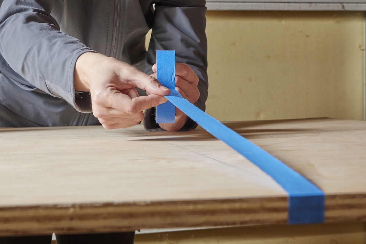 Woman uses blue painter's tape to tape a cut line on plywood.