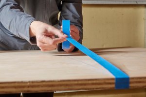 Woman uses blue painter's tape to tape a cut line on plywood.