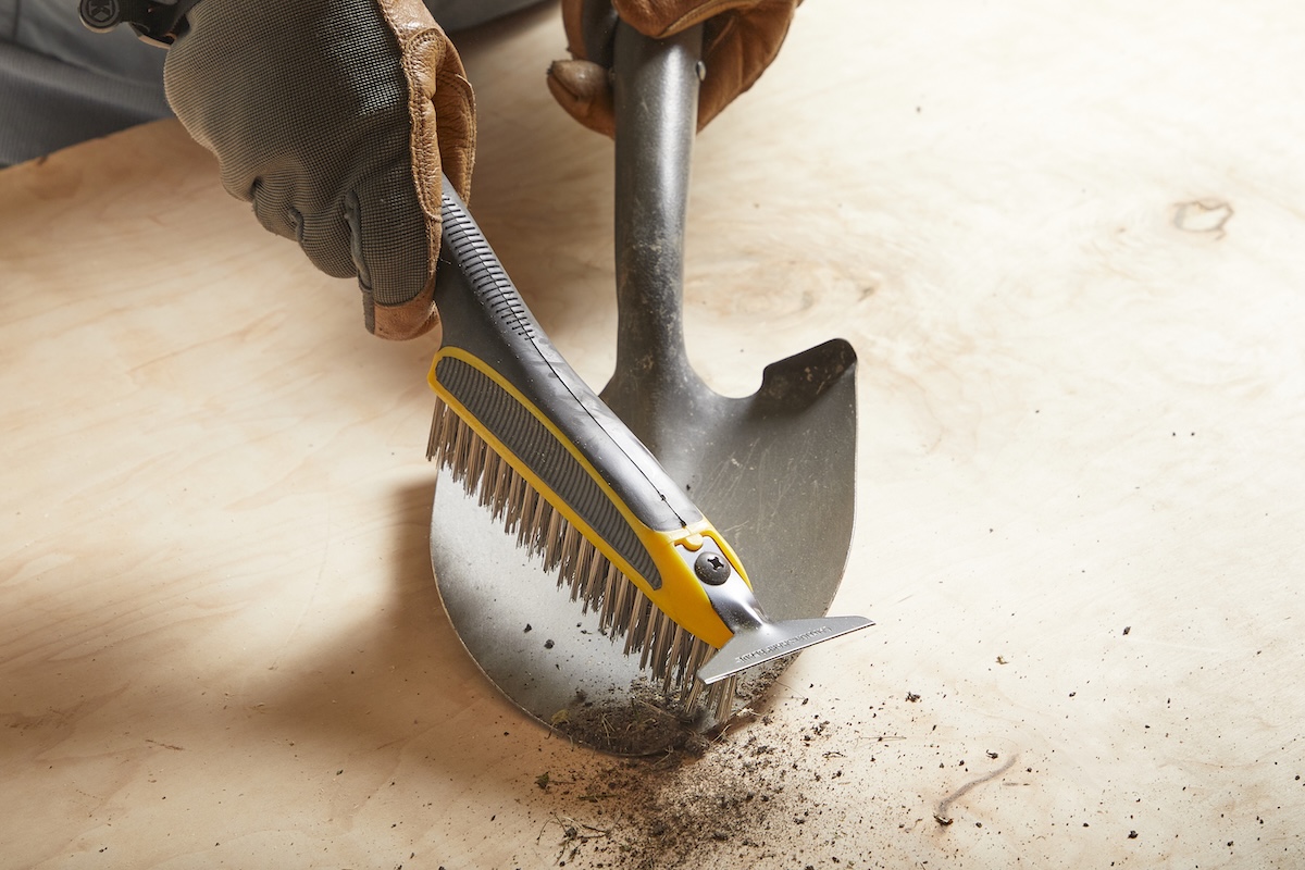 Person wearing work gloves scrubs a shovel with a wire brush.