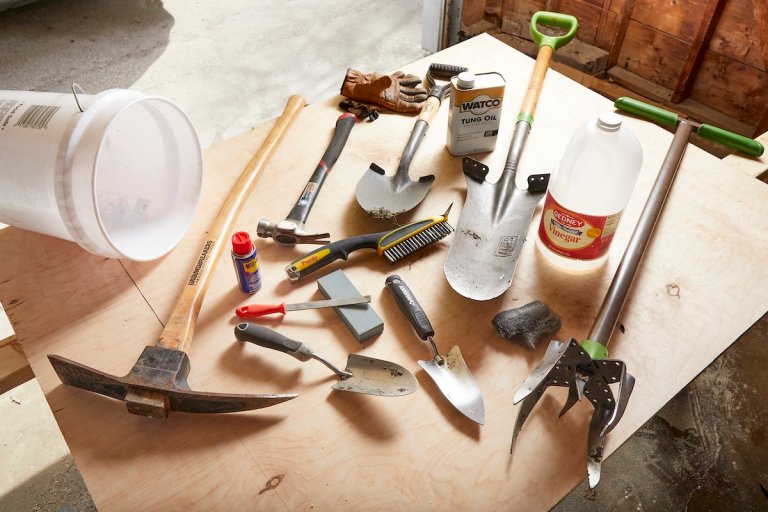 Many garden tools like shovels and trowels laid out on a wooden workshop table in the sunlight.
