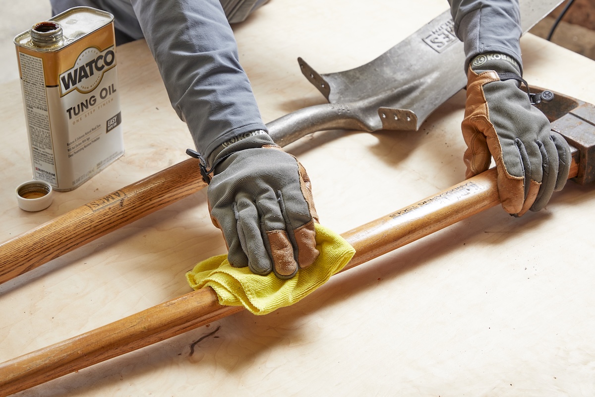 A woman in work gloves rubs the wooden handles of garden tools with tung oil.