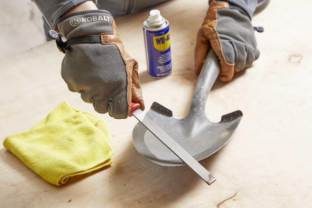 Woman uses file on shovel head, WD-40 in background.
