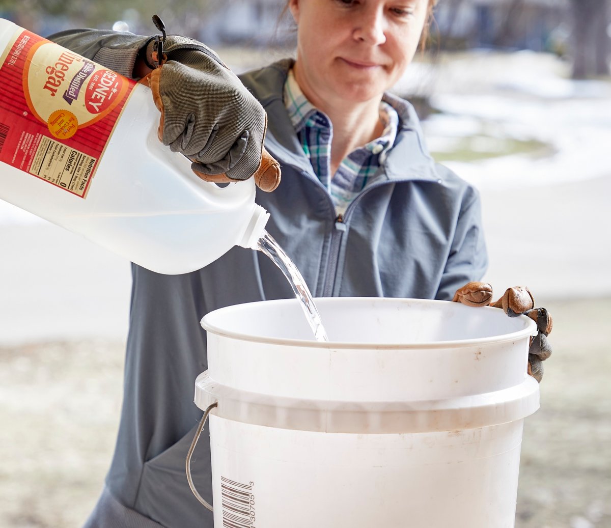 A woman pours vinegar into a white 5-gallon bucket outside.