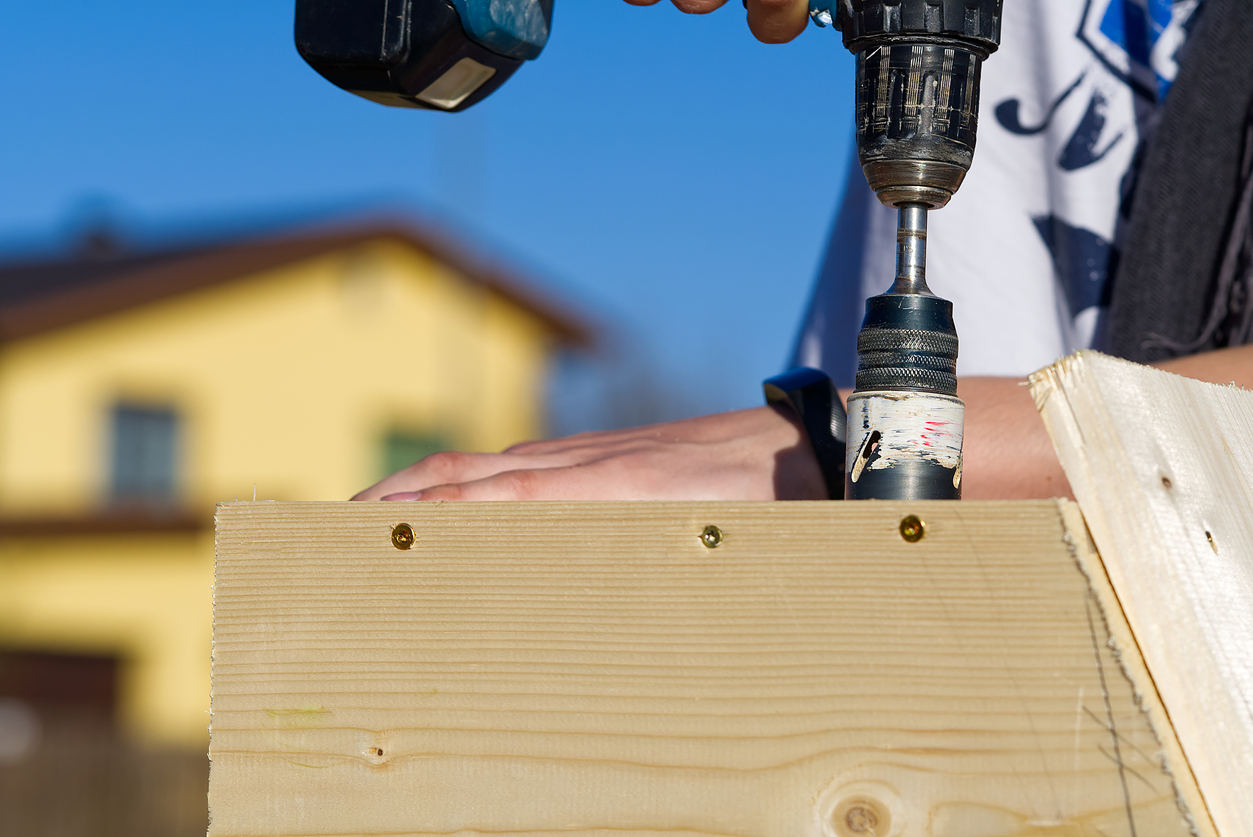 Using a hole saw on drill to drill hole into wooden house structure with a yellow house and blue ski in the background.