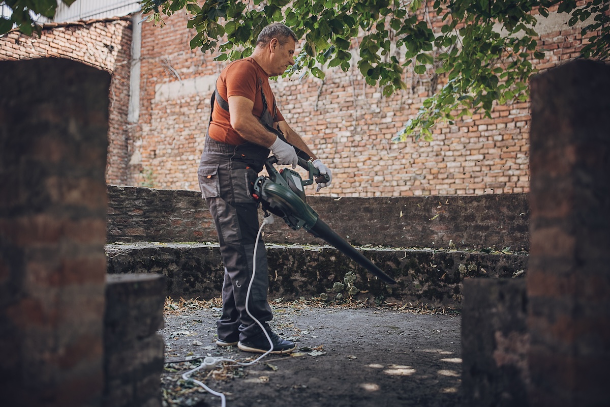 A man uses a leaf blower to push the leaves toward a stone fence.