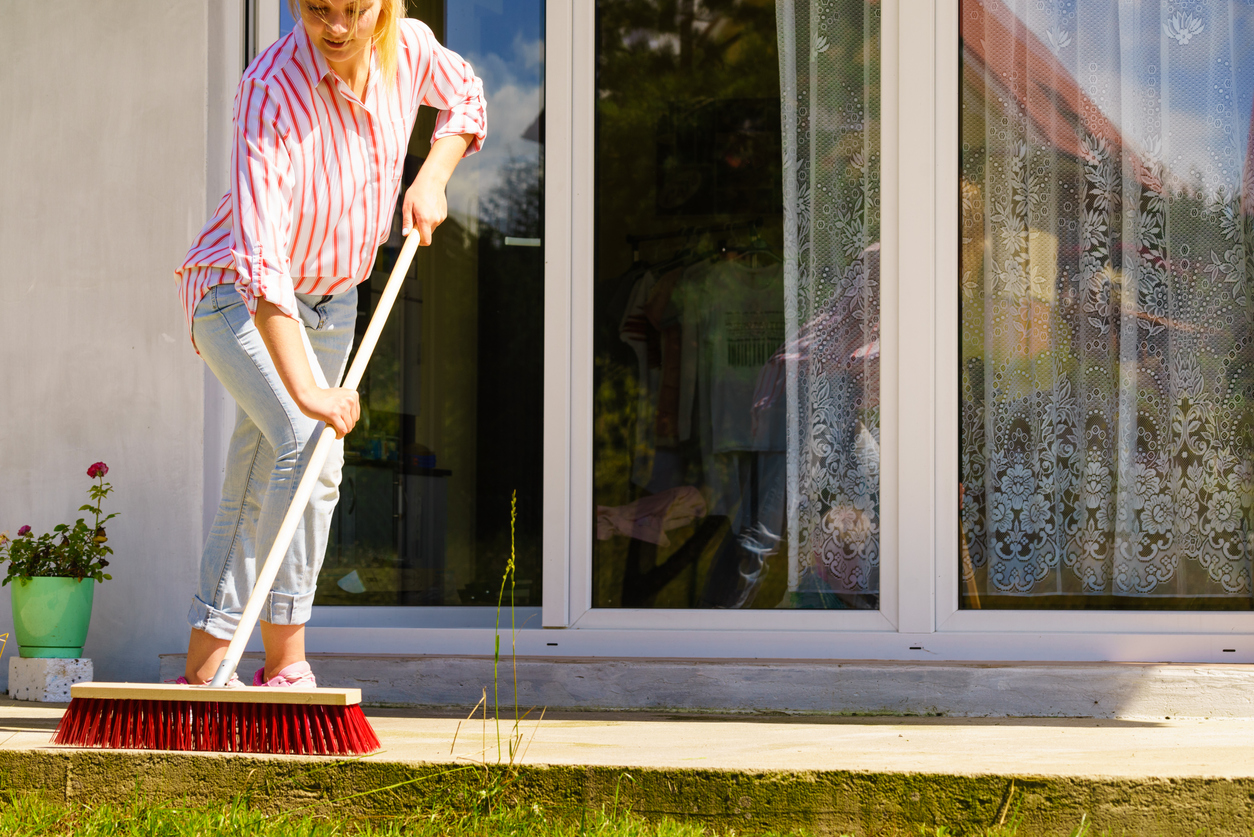 woman in pink and white striped shirt sweeping patio with red bristled broom in front of patio door of house leading to living room