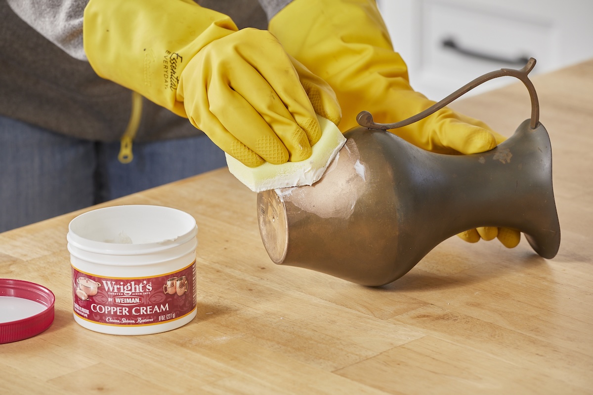 Person wearing yellow rubber gloves applies metal cream to tarnished brass pitcher.