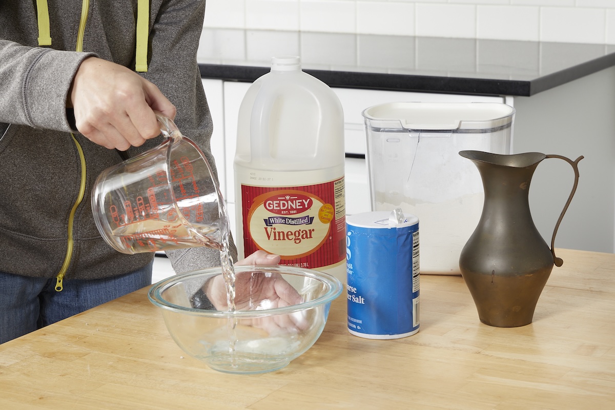 Woman combines vinegar, salt, and flour in a glass bowl, a tarnished brass pitcher nearby.