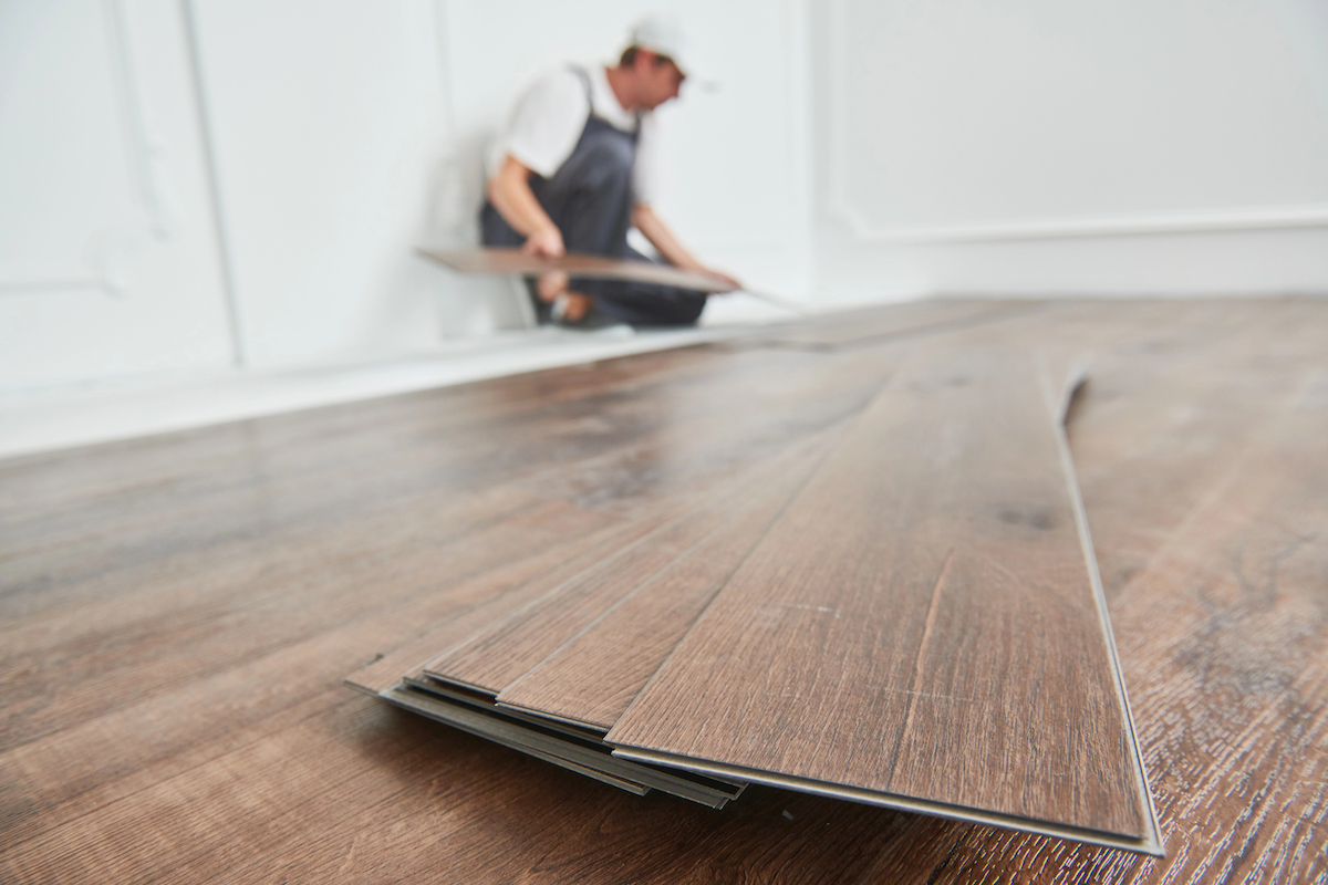 In foreground, pile of vinyl plank flooring over a finished floor. A worker in the background.