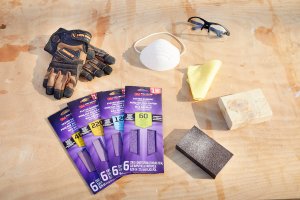 Sanding materials such as sandpaper, mask, eye protection, and block sander laid out on a wood table.