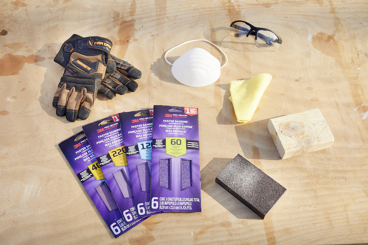 Sanding materials such as sandpaper, mask, eye protection, and block sander laid out on a wood table.