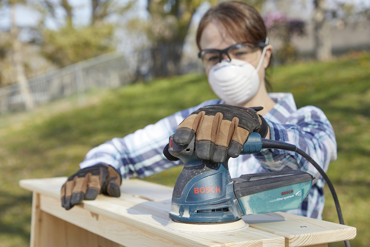 Woman wearing dust mask uses a sander to sand a wood drawer.