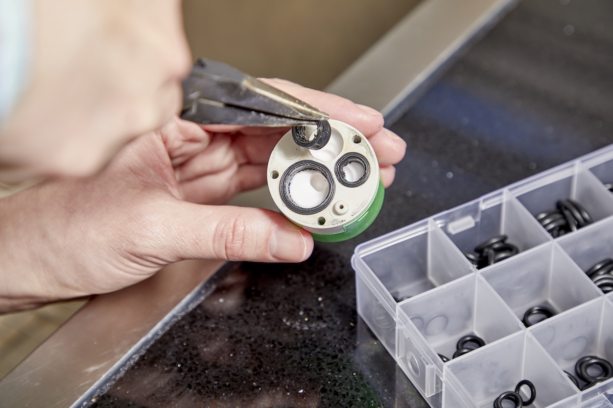 Person holds faucet cartridge in one hand, needle-nose pliers removing a cartridge O-ring with the other.