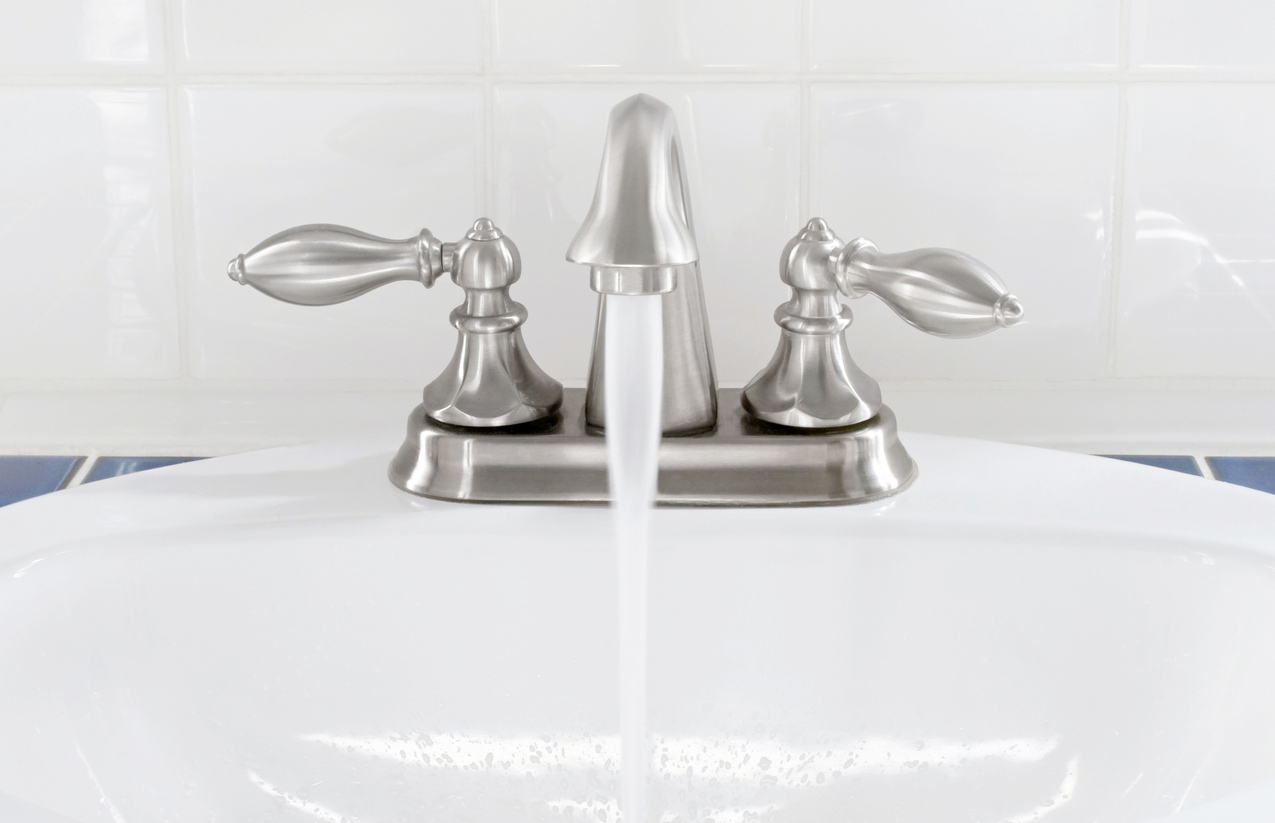 Brushed nickel faucet & handles with running water into white sink.