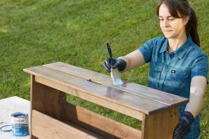 Person in blue blouse applies a wood sealant onto a freshly stain wooden shelf with a paint brush.