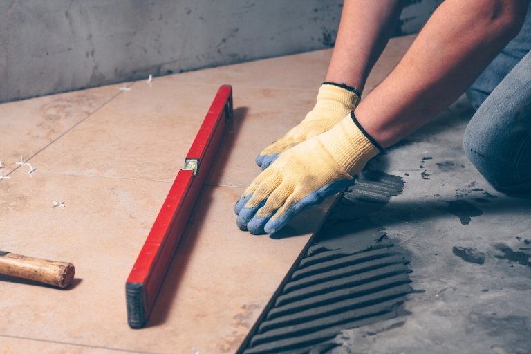 Worker's hands in gloves press the tile to the floor as part of tile installation. Level sits nearby.