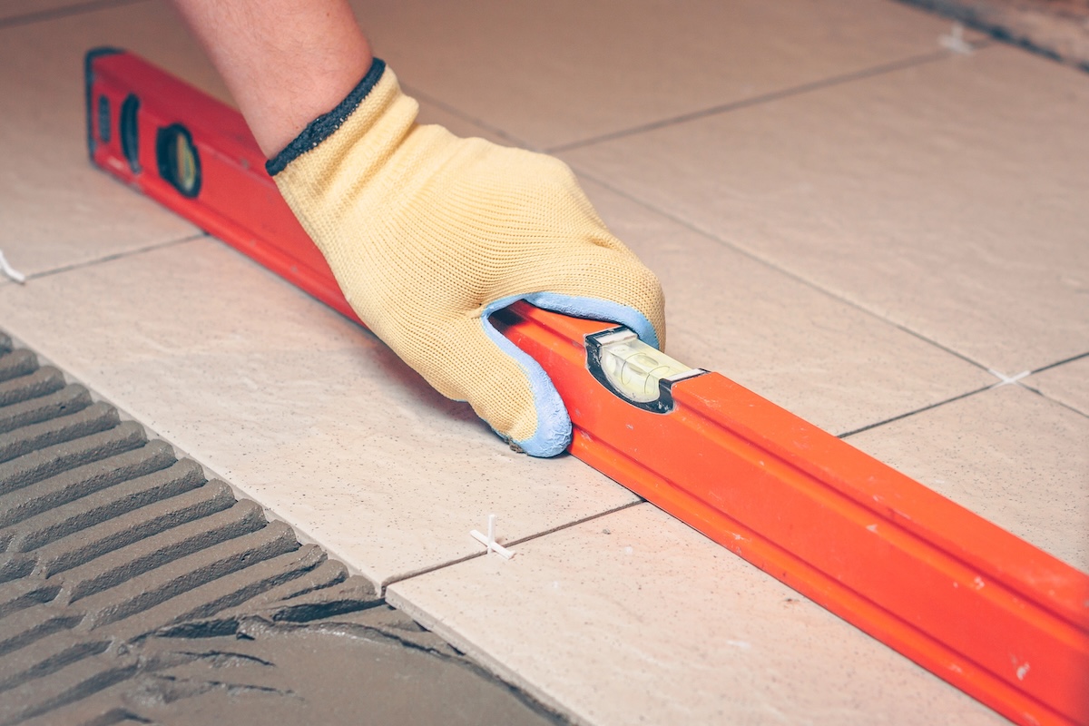 Person wearing yellow work gloves uses a level to check tile floor installation.