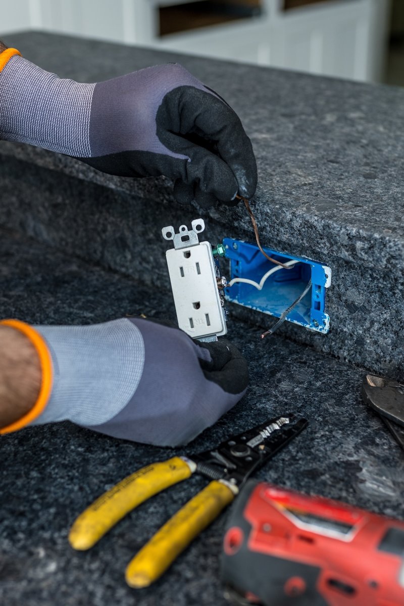 Man wearing work gloves installs GFCI outlet on kitchen island top with dark-colored granite. 