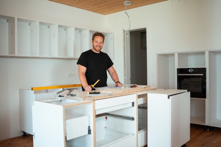 A smiling man with tools assembles a modern kitchen interior, dressed casually for hands-on work.