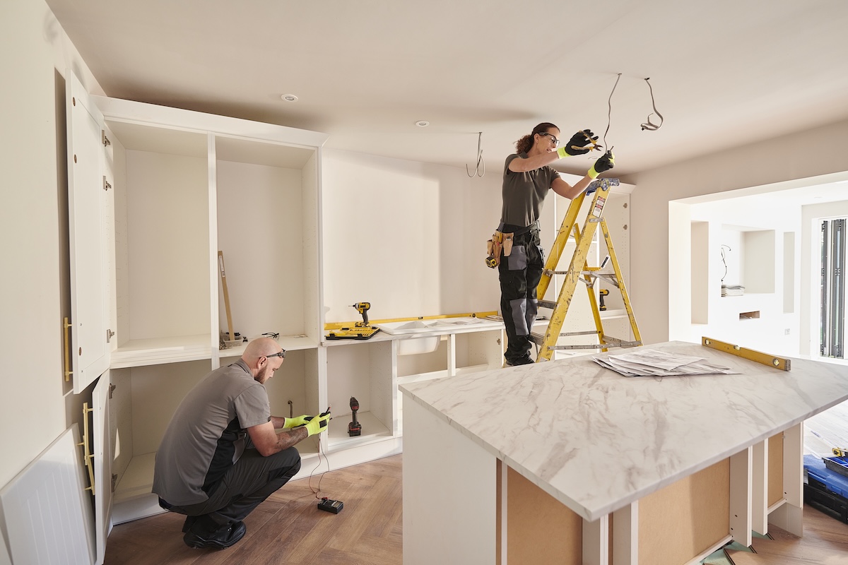 Female electrician on a ladder wiring an overlead light in a kitchen remodel while a male contractor works near cabinets without doors.