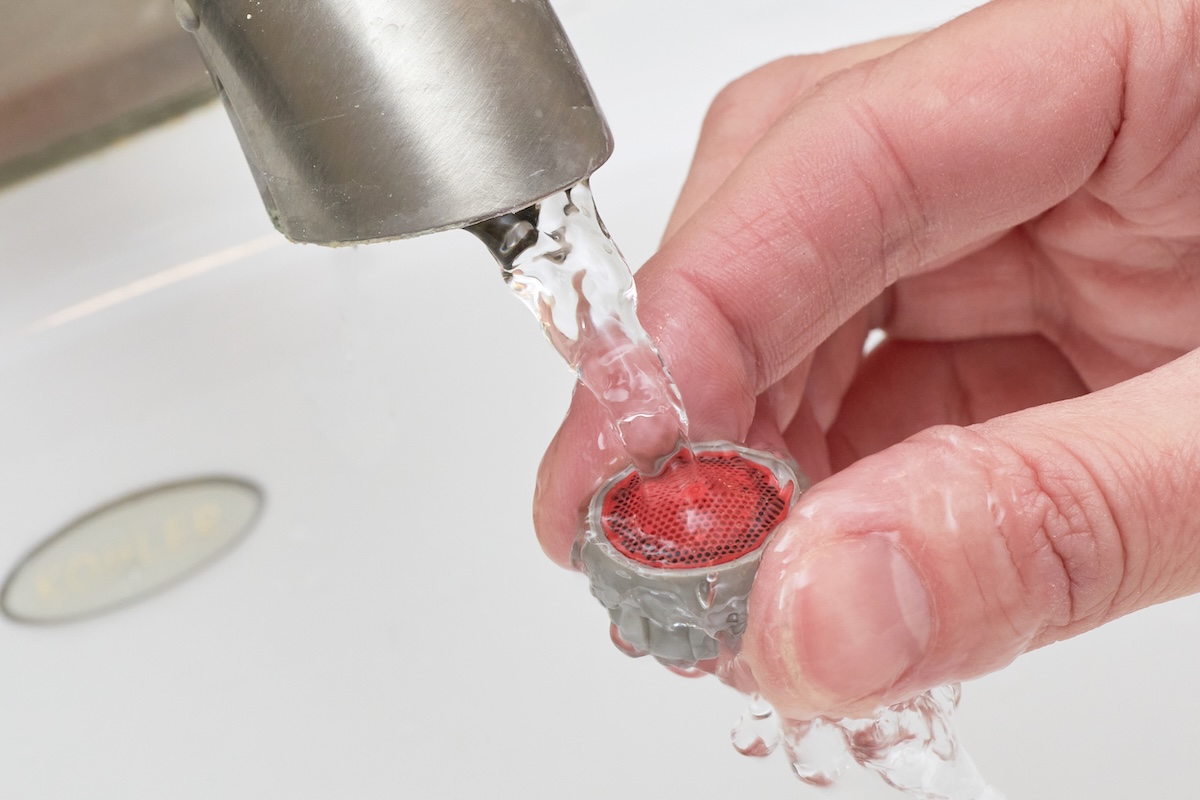 A hand holds a faucet aerator under a tap to clean it with water.
