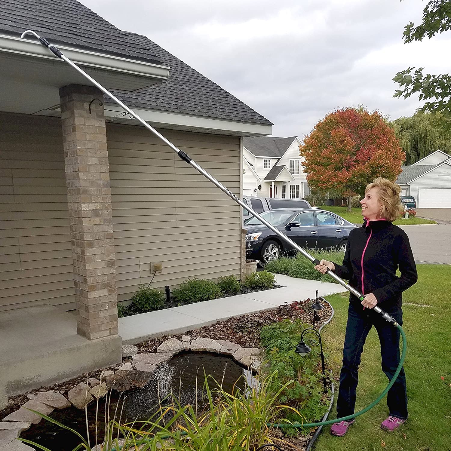 Woman in a black tracksuit using a gutter extension pole to clean her home's rain gutters.