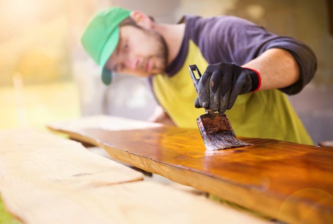 Man with green baseball cap varnishes a plank of wood.