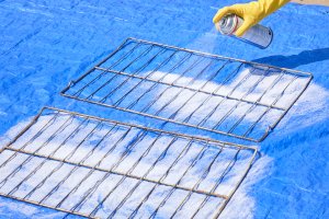 Woman sprays oven racks with oven cleaner on a blue plastic tarp.