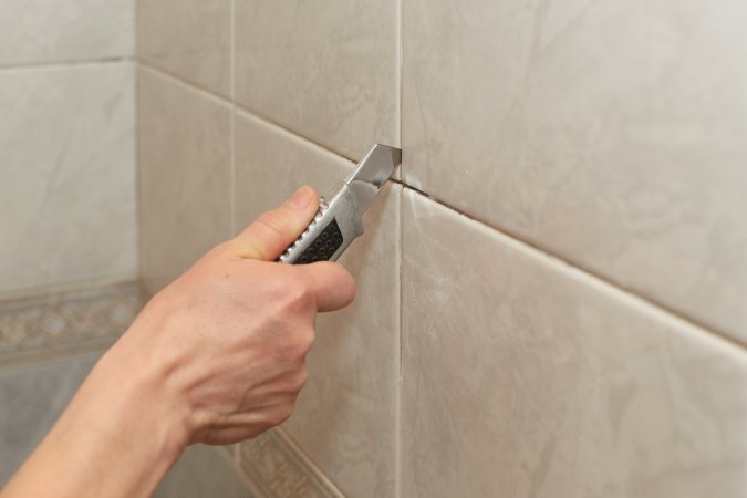Male hands with knife removing the old grout from beige tile.