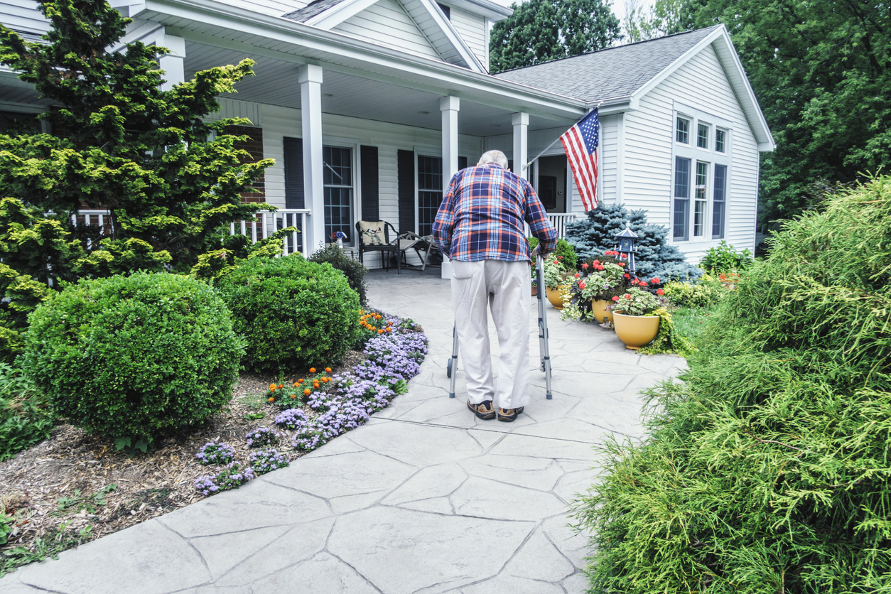 senior man uses walker to walk up pathway to front door of suburban home