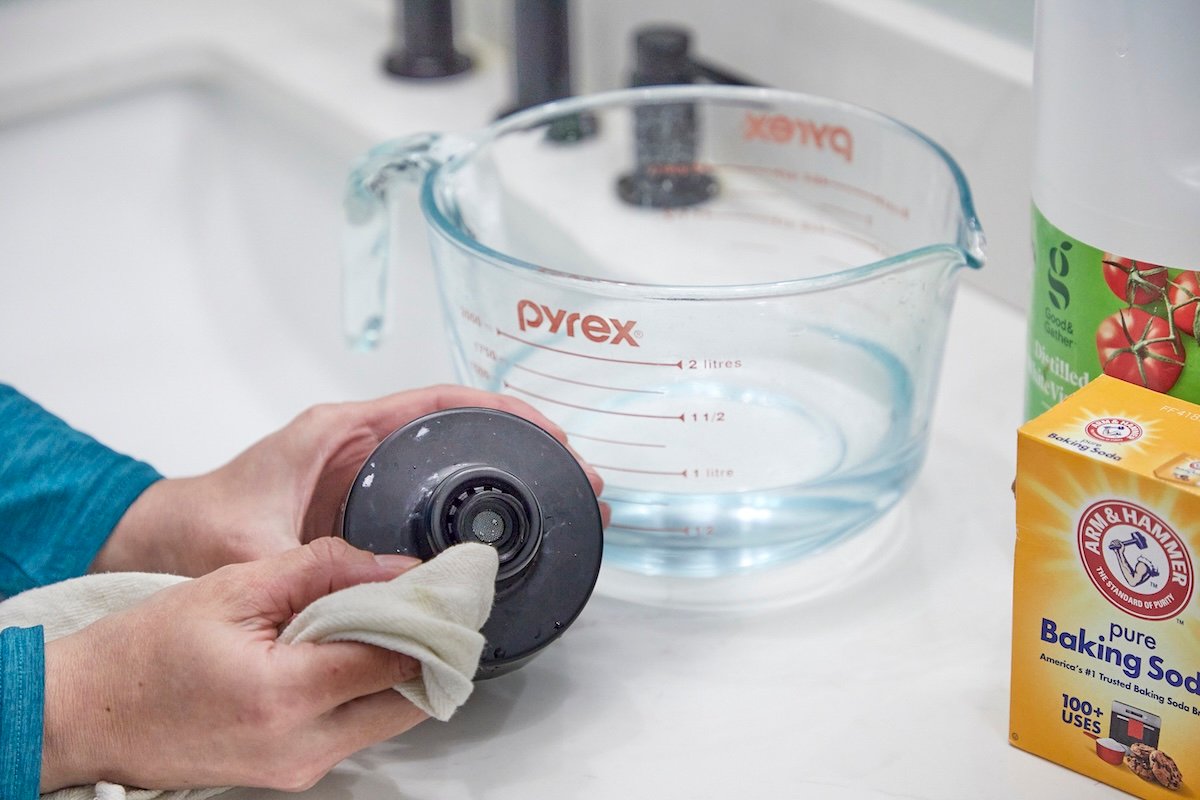A woman cleans the underside of a separate shower head with a cloth, next to the bathroom sink.