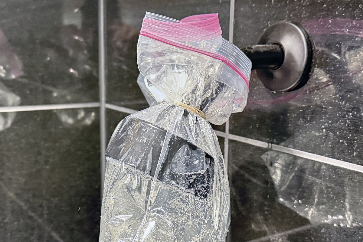 A ziploc bag filled with a vinegar solution is tied around a shower head attached to a grey tile wall, held in place with a rubber band.