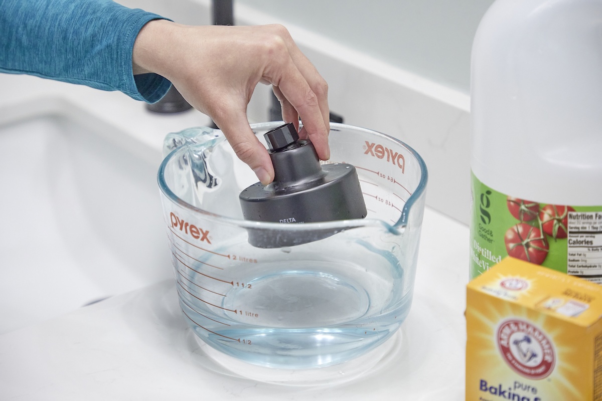 Woman soaks a shower head in a glass measuring cup with a baking soda and vinegar solution.