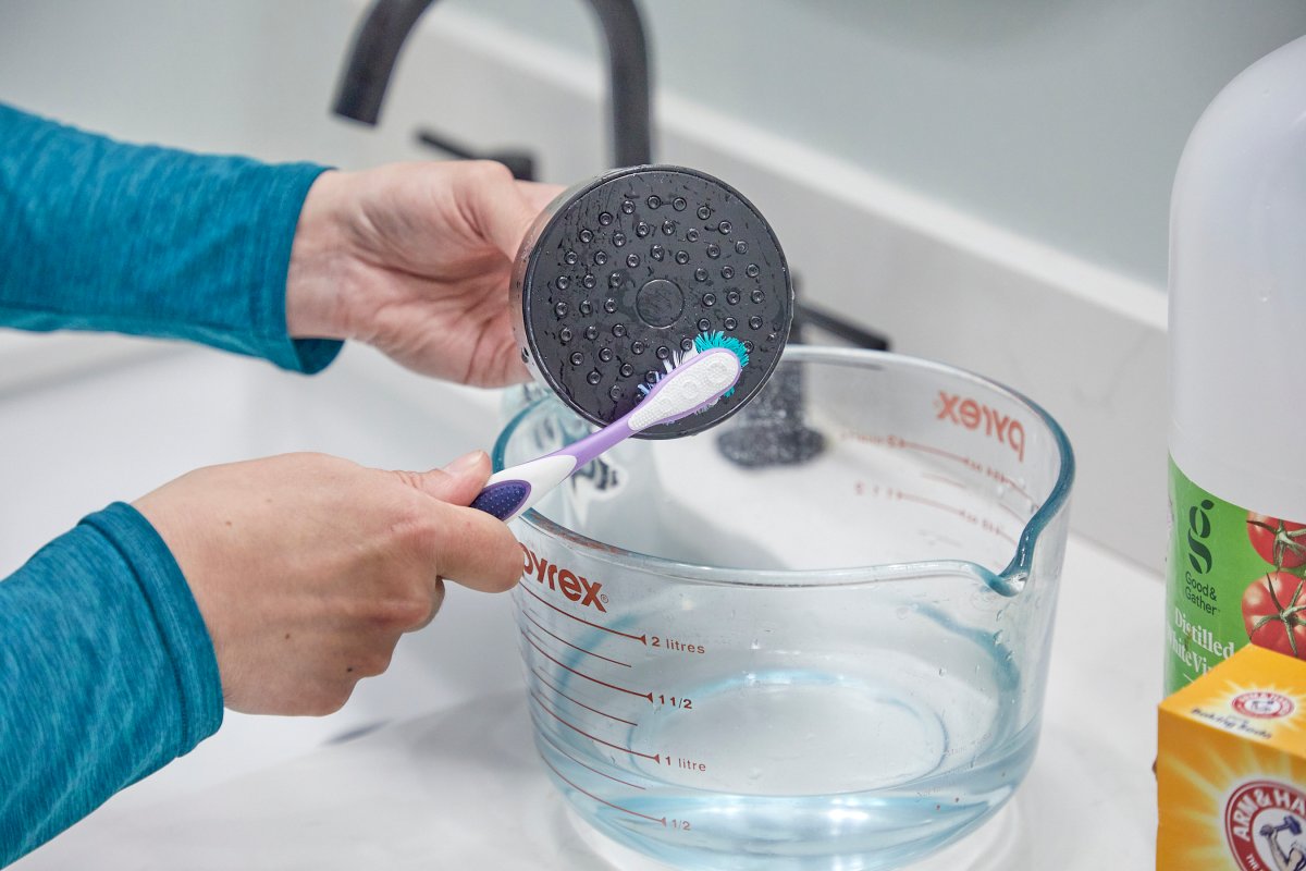 Woman cleans shower head with toothbrush over oversized cup.