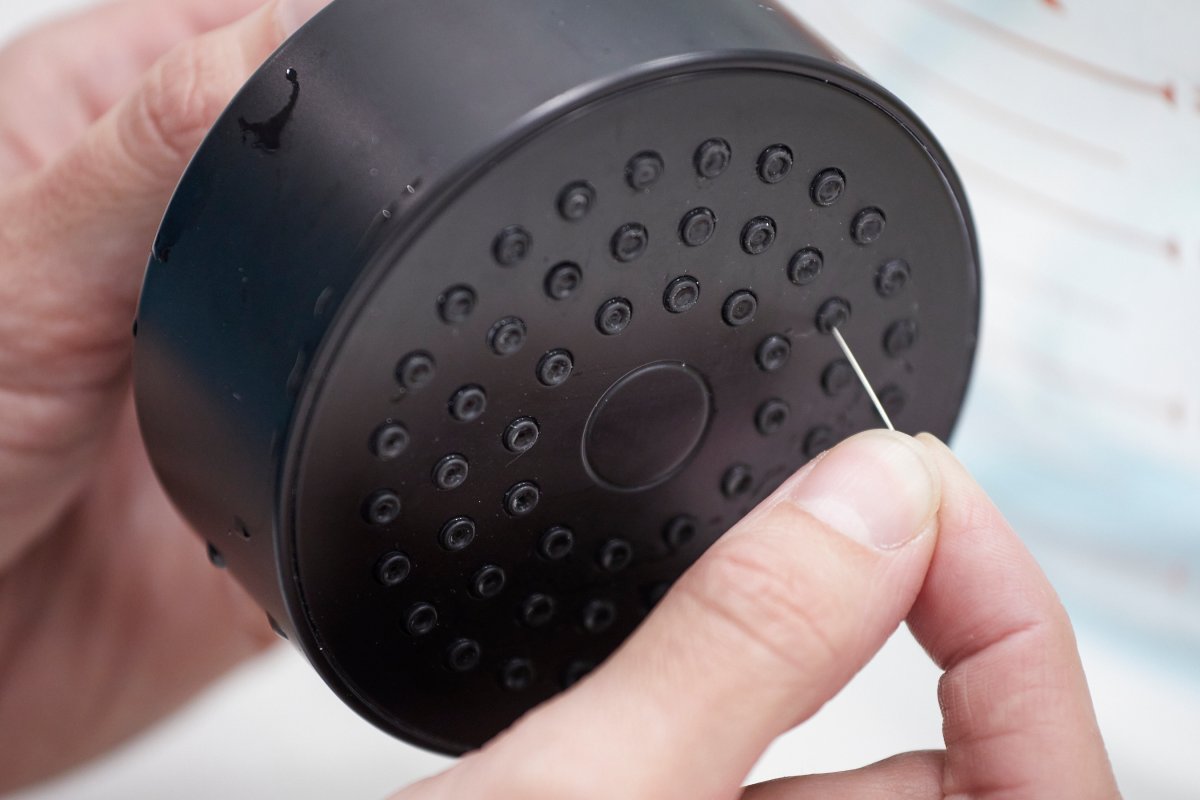 Woman holds black shower head in hand, using needle to clean spray nozzle.