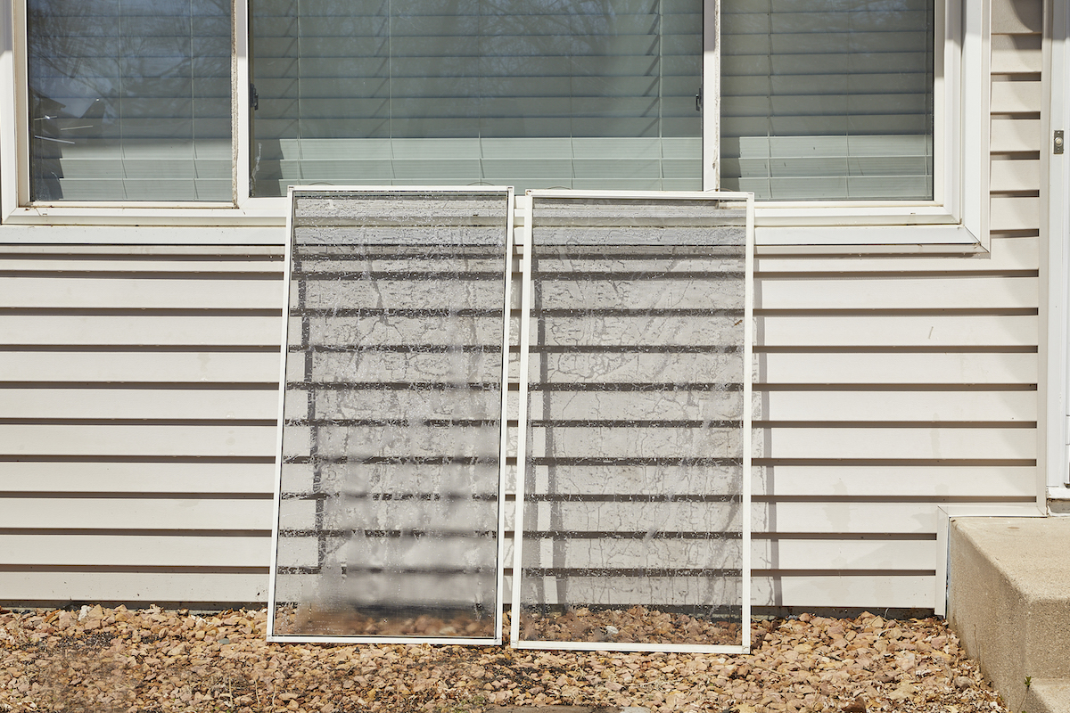 Two window screens propped up under a window against a house with beige vinyl siding.