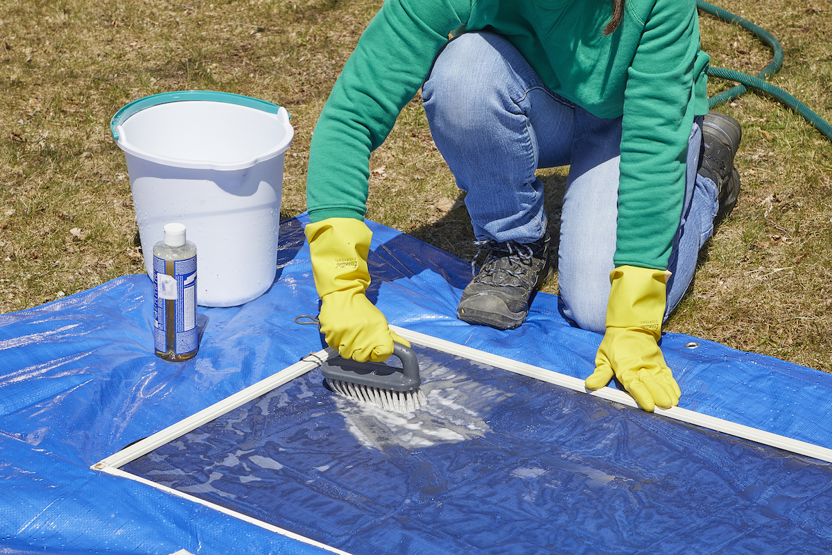 Woman wearing rubber gloves scrubs window screens lying on a blue tarp.