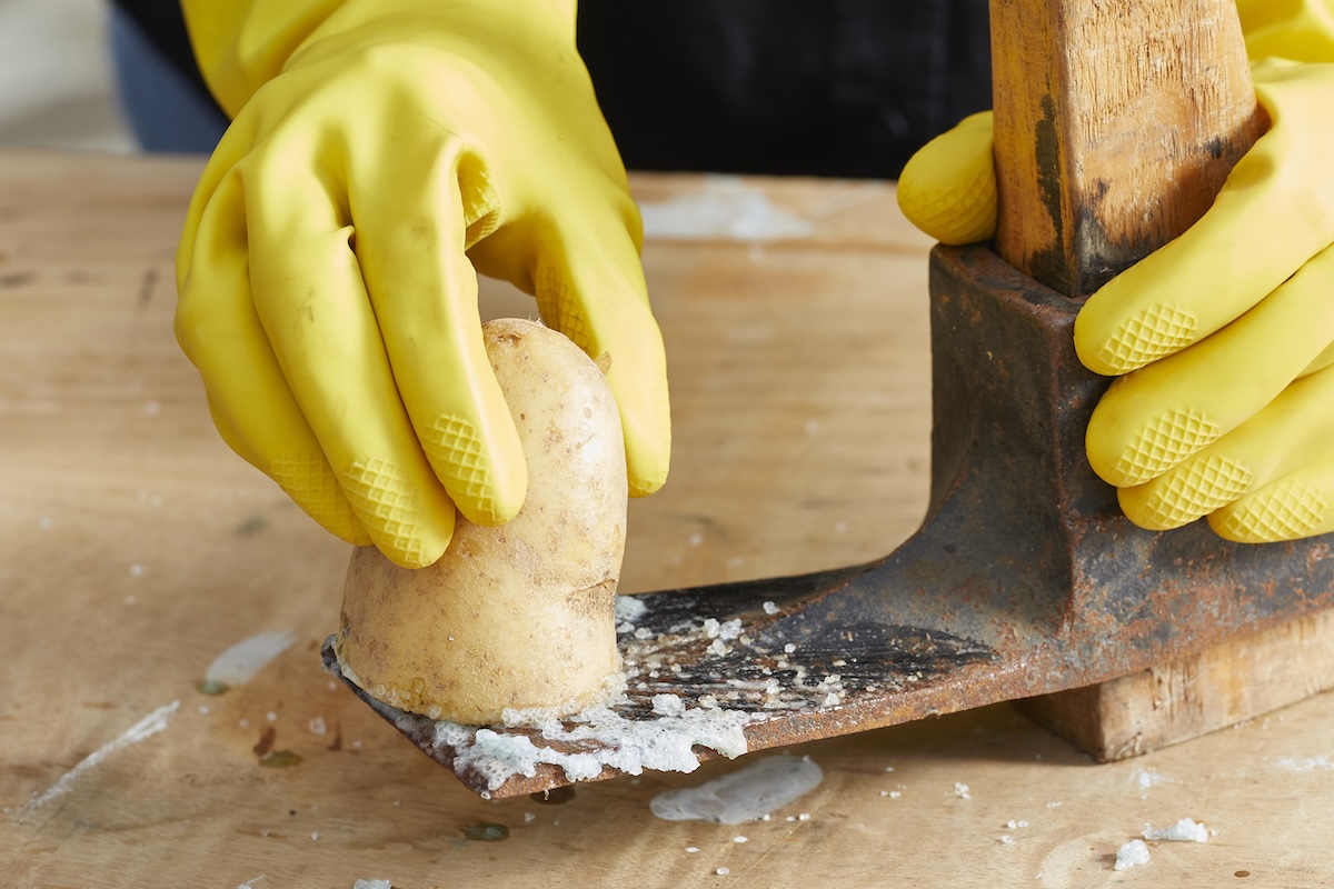 Person wearing rubber gloves rubs potato on a rusty ax blade.