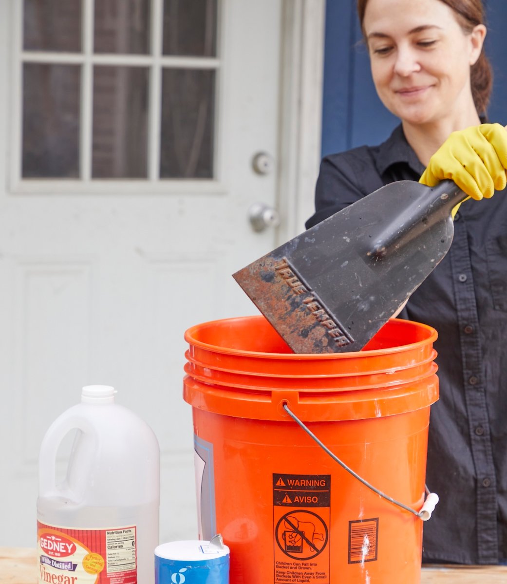 Woman dunks rusty tool into orange 5-gallon bucket filled with vinegar, salt, and water.