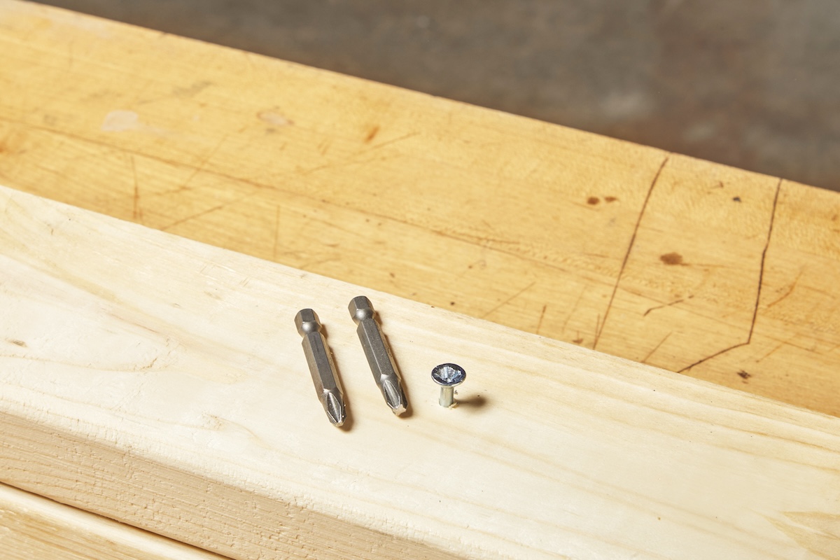 Two different sizes of drill bits laying next to a stripped screw in a wood stud on a home workbench.
