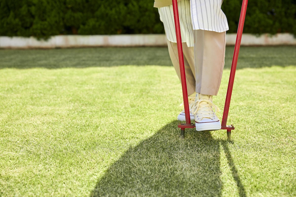 A woman wearing yellow sneakers uses a core aerator on her lawn.