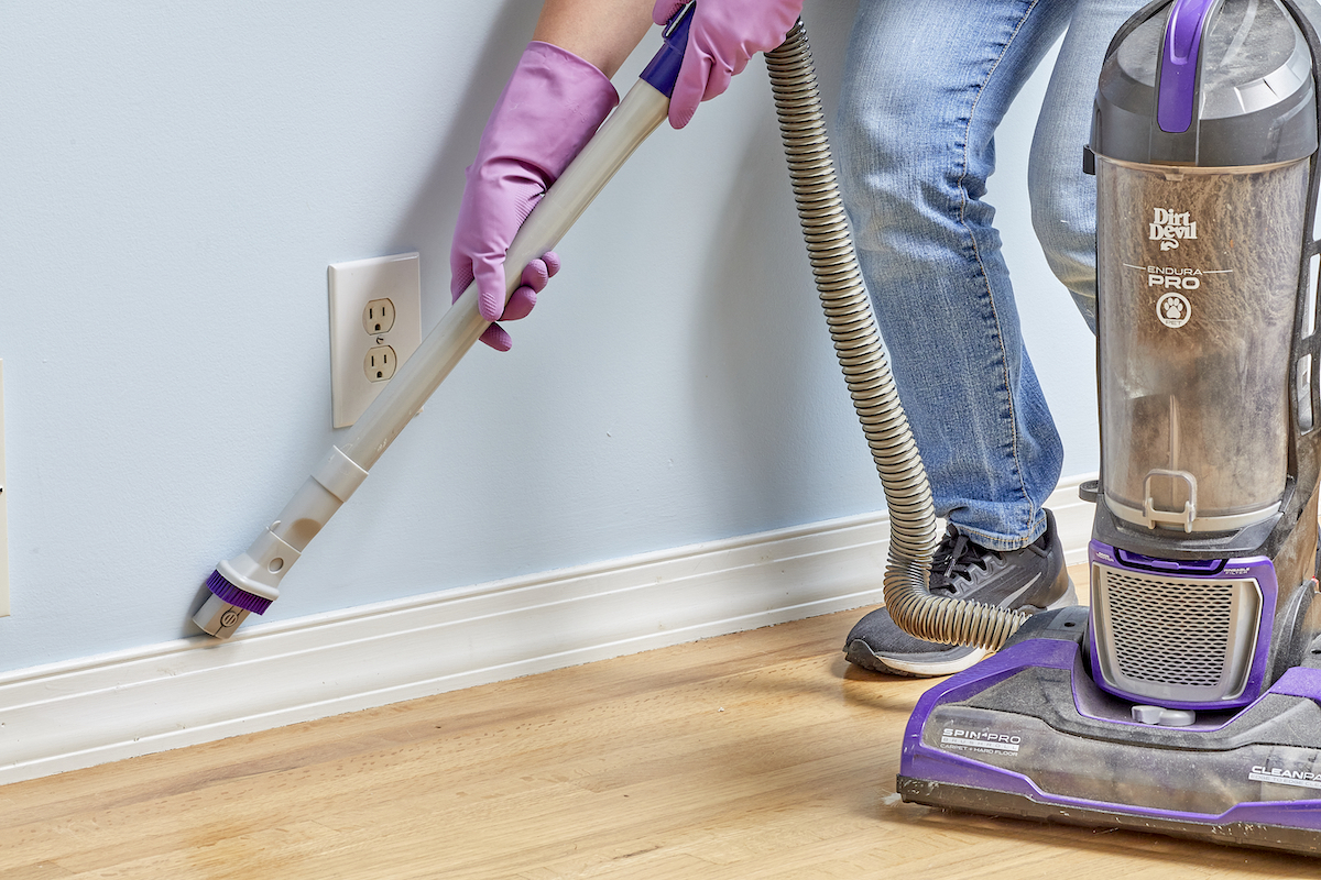 Woman vacuums white baseboards using a vacuum attachment.