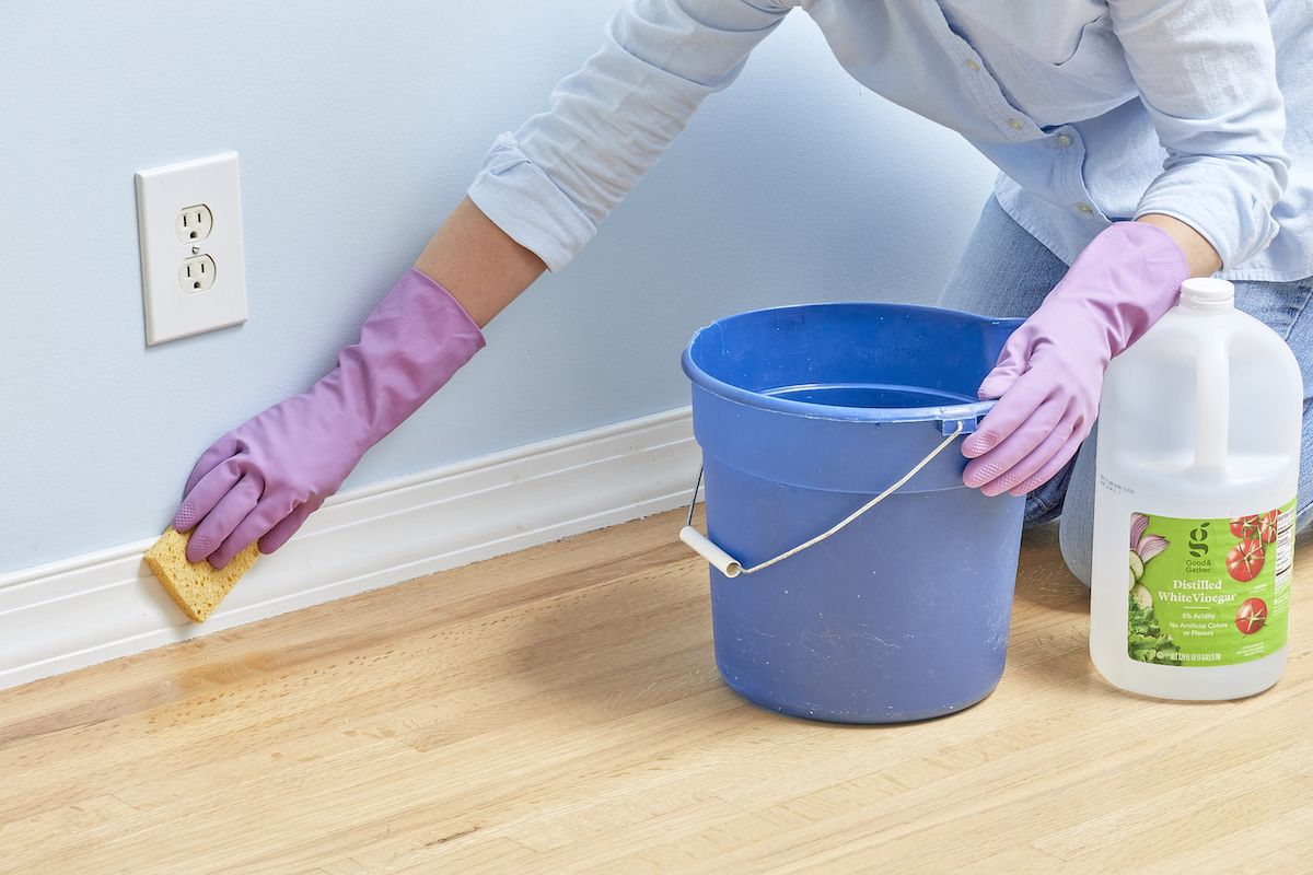 Woman wipes baseboards with a sponge, a blue bucket and a bottle of vinegar nearby.